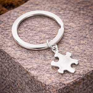 May include: A silver-colored keychain with a puzzle piece charm. The keychain ring is round and attached to a small, shiny puzzle piece. The items are displayed on a textured, brown surface.