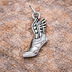 May include: A silver-colored charm in the shape of a running shoe with wings. The shoe has detailed lines and a loop at the top for attaching to a necklace or bracelet. The wings are black and white.