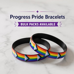 May include: Two black bracelets with a rainbow Progress Pride flag design. The bracelets have a black interior and a colorful exterior with the Progress Pride flag. The text "Progress Pride Bracelets" and "Bulk Packs Available" is visible.