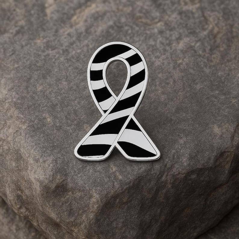 May include: A silver-toned ribbon pin with a black and white striped design. The pin is in the shape of a looped ribbon, with a polished silver border. The pin is set against a gray stone background.