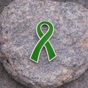 May include: A green ribbon pin with a white border, resting on a gray stone surface. The ribbon is a symbol of awareness and support. The pin is a simple yet meaningful accessory.