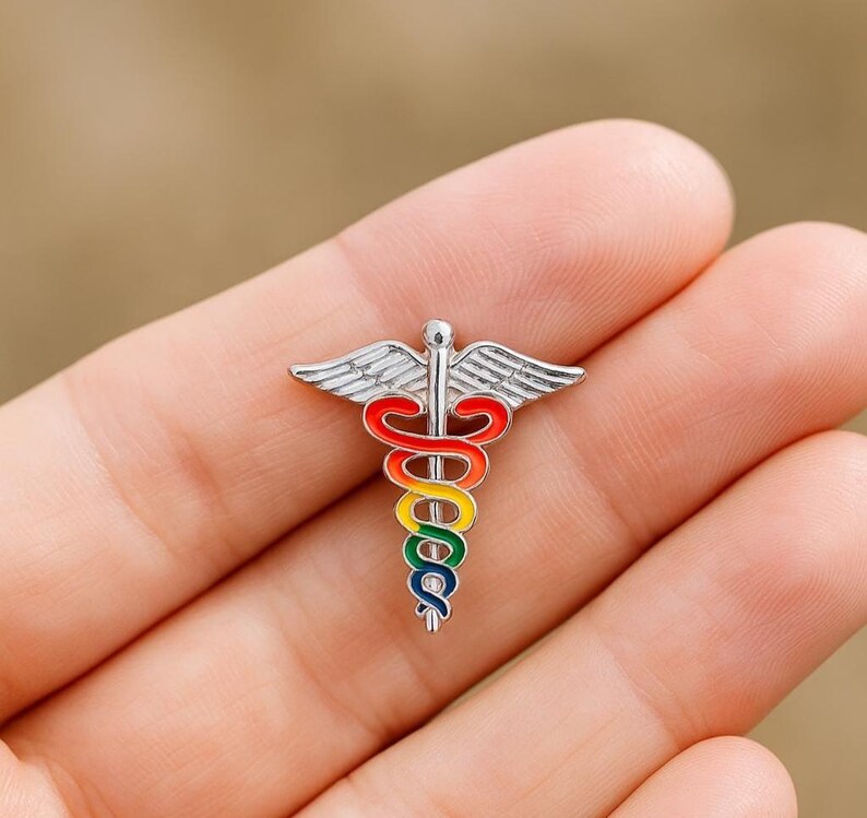 Peut inclure: &Eacute;pingle Caduc&eacute;e argent&eacute;e avec un serpent et des ailes aux couleurs de l'arc-en-ciel. Ce symbole m&eacute;dical est un accessoire color&eacute;, id&eacute;al pour les professionnels de la sant&eacute;. L'&eacute;pingle est petite et d&eacute;taill&eacute;e, parfaite pour une veste.
