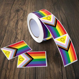 May include: A roll of rainbow-colored stickers with a Pride flag design, featuring a triangle with a circle and stripes. Two individual stickers are also visible, all on a wooden surface.