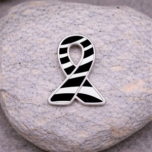 May include: A black and white striped ribbon pin with a silver border. The pin is in the shape of a ribbon, with a loop at the top and two ends that cross at the bottom. The pin is set against a gray stone background.
