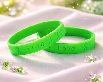 May include: Two bright green silicone wristbands with the word "Love" and a ribbon symbol embossed on them. The bands are resting on a white, silky surface. The background has soft, blurred green and white accents.