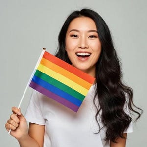 May include: A rainbow flag, featuring stripes of red, orange, yellow, green, blue, and purple, is held up. The flag is on a white pole. The person holding the flag is smiling and wearing a white t-shirt.