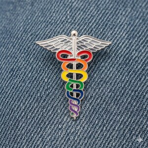 Peut inclure: &Eacute;pinglette en m&eacute;tal argent&eacute; repr&eacute;sentant le symbole du Caduc&eacute;e, avec un serpent aux couleurs de l'arc-en-ciel enroul&eacute; autour d'un b&acirc;ton et des ailes. L'&eacute;pinglette est pos&eacute;e sur un fond en denim bleu.