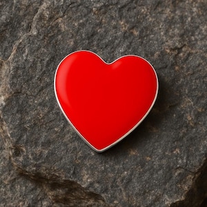 May include: A vibrant red heart-shaped pendant with a silver-colored border. The heart is set against a textured, dark gray stone surface, creating a striking contrast. The pendant's smooth, glossy finish catches the light.