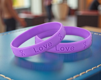 May include: Two lavender rubber bracelets with the word "Love" and a ribbon symbol embossed on them. The bracelets are resting on a blue surface. The bracelets are a symbol of support and awareness.