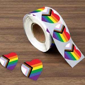 May include: A roll of heart-shaped stickers with a rainbow design and the Progress Pride flag. Two individual stickers are next to the roll. The stickers are on a wooden surface.