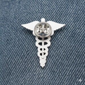 Peut inclure: &Eacute;pinglette argent&eacute;e repr&eacute;sentant un caduc&eacute;e m&eacute;dical, avec des ailes et un b&acirc;ton, sur un fond en denim. La broche pr&eacute;sente un design d&eacute;taill&eacute;, avec un b&acirc;ton central et deux serpents.