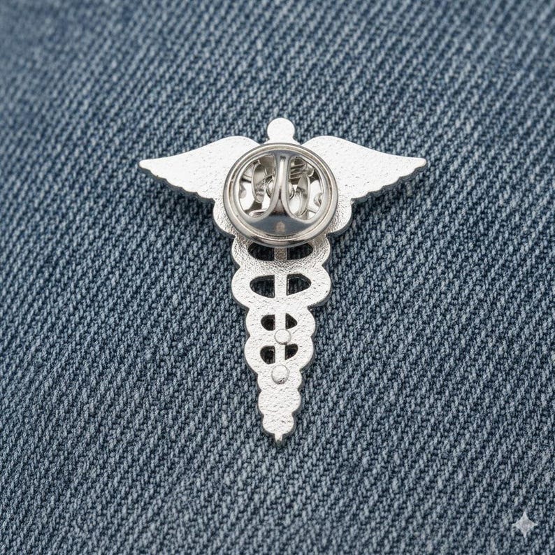 Peut inclure: &Eacute;pinglette argent&eacute;e repr&eacute;sentant un caduc&eacute;e m&eacute;dical, avec des ailes et un b&acirc;ton, sur un fond en denim. La broche pr&eacute;sente un design d&eacute;taill&eacute;, avec un b&acirc;ton central et deux serpents.