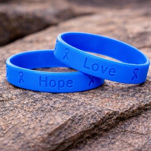 May include: Two blue silicone wristbands with the words "Hope" and "Love" embossed on them. Each band features a ribbon symbol. The wristbands are resting on a textured, brown stone surface.