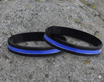 May include: Two black silicone wristbands with a vibrant blue stripe. The bands are circular and lie on a textured gray stone surface. The blue stripe is centered on each band, creating a simple yet striking design.