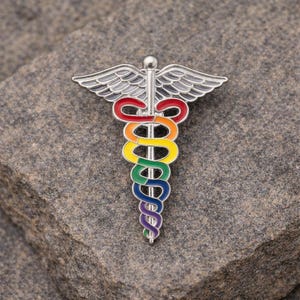 Peut inclure: Une &eacute;pingle en m&eacute;tal argent&eacute; en forme de caduc&eacute;e, avec un serpent et des ailes aux couleurs de l'arc-en-ciel. L'&eacute;pingle est pos&eacute;e sur un fond de pierre grise textur&eacute;e. Les couleurs du serpent sont rouge, orange, jaune, vert, bleu et violet.