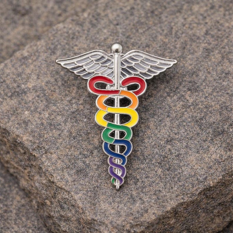 Peut inclure: Une &eacute;pingle en m&eacute;tal argent&eacute; en forme de caduc&eacute;e, avec un serpent et des ailes aux couleurs de l'arc-en-ciel. L'&eacute;pingle est pos&eacute;e sur un fond de pierre grise textur&eacute;e. Les couleurs du serpent sont rouge, orange, jaune, vert, bleu et violet.