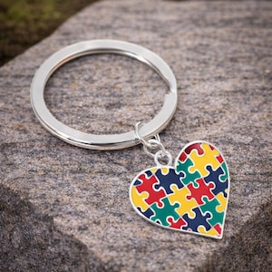 May include: A silver-colored keychain with a heart-shaped pendant. The pendant features a colorful puzzle piece design in red, yellow, green, and blue. The keychain is attached to a large, round key ring.