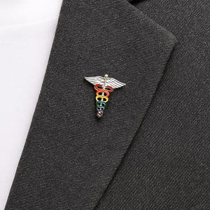 Peut inclure: &Eacute;pinglette argent&eacute;e en forme de caduc&eacute;e, avec un b&acirc;ton et des ailes aux couleurs de l'arc-en-ciel. L'&eacute;pinglette est fix&eacute;e &agrave; un blazer gris fonc&eacute;, avec une chemise blanche en dessous.