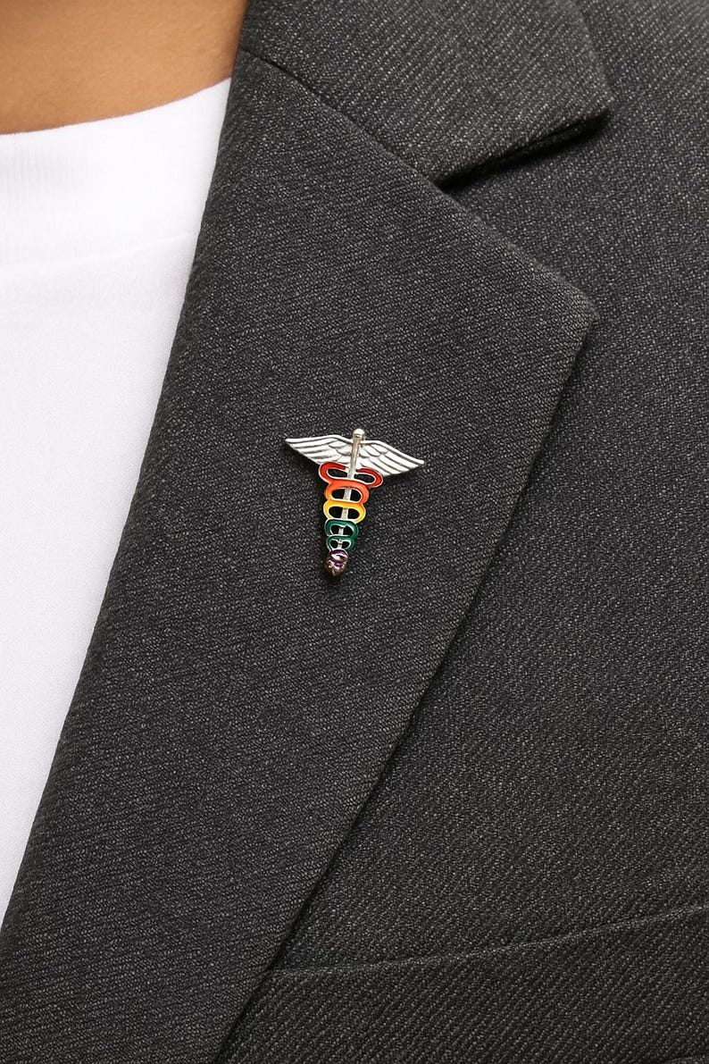 Peut inclure: &Eacute;pinglette argent&eacute;e en forme de caduc&eacute;e, avec un b&acirc;ton et des ailes aux couleurs de l'arc-en-ciel. L'&eacute;pinglette est fix&eacute;e &agrave; un blazer gris fonc&eacute;, avec une chemise blanche en dessous.