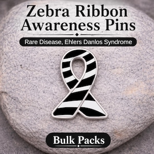 May include: A zebra ribbon awareness pin with black and white stripes, set against a gray stone background. The pin is for rare disease and Ehlers Danlos Syndrome awareness. Text on the image reads "Zebra Ribbon Awareness Pins", "Rare Disease, Ehlers Danlos Syndrome", and "Bulk Packs".