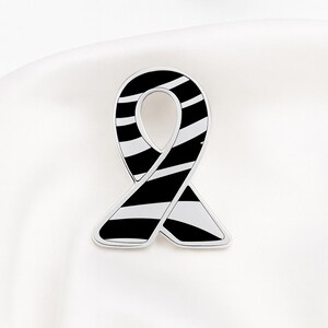 May include: A silver-toned ribbon-shaped pin with a black and white striped pattern. The pin is set against a white background, highlighting its design. The ribbon design is a symbol of awareness.
