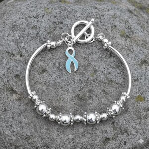 May include: Silver bracelet featuring a light blue ribbon charm. The bracelet has a toggle clasp and is adorned with silver beads. The charm is a light blue ribbon, a symbol of awareness. The bracelet is displayed on a gray stone surface.