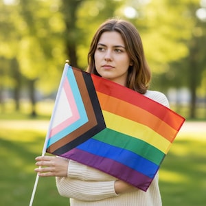May include: A rainbow flag with a progressive design, featuring stripes of red, orange, yellow, green, blue, and purple, along with a chevron of pink, light blue, brown, black, and white. The flag is held against a blurred green background.