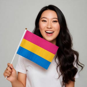 May include: A small flag with a pink, yellow, and blue horizontal stripe design. The flag is held by a person wearing a white t-shirt. The flag is on a white pole with a gold tip.