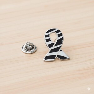 May include: A silver-colored ribbon pin with black and white stripes. The pin is in the shape of a ribbon, with a pin back on the left side. The ribbon has a glossy finish and is set against a light wooden surface.