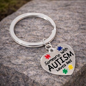 May include: A silver heart-shaped keychain with the words "Someone With AUTISM Loves Me!" and colorful puzzle piece accents. The keychain is attached to a silver ring.