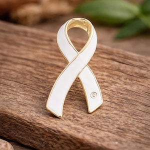 May include: A white ribbon pin with a gold-colored border and a small, round accent. The pin is set against a wooden background, highlighting its design and color contrast. The ribbon shape is a symbol of awareness.