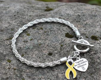May include: Silver-toned bracelet with a braided design. A yellow ribbon charm and a heart-shaped charm with the text "Together we can make a difference" hang from the bracelet. The bracelet is resting on a gray stone surface.