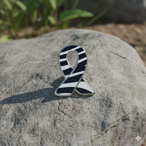 May include: A ribbon-shaped enamel pin with a black and white zebra stripe pattern. The pin has a silver-colored border and is set against a gray stone background. The pin is a symbol of awareness.