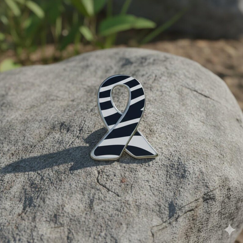May include: A ribbon-shaped enamel pin with a black and white zebra stripe pattern. The pin has a silver-colored border and is set against a gray stone background. The pin is a symbol of awareness.
