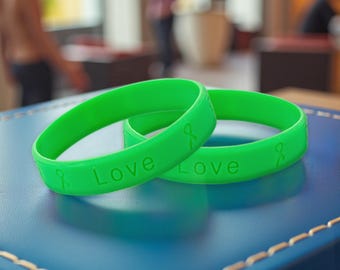 May include: Two bright green silicone wristbands with the word "Love" and a ribbon symbol embossed on them. The wristbands are resting on a blue surface. The wristbands are a symbol of support and awareness.