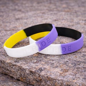 May include: Two silicone wristbands with the word "PRIDE" embossed on them. One band is white, yellow, and black. The other is white, purple, and black. The bands are resting on a rough, gray stone surface.