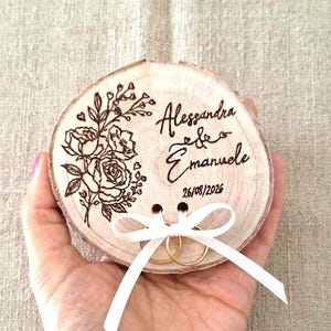 Può includere: Un portafedi circolare in fetta di legno con un disegno floreale pirografato sul lato sinistro e i nomi incisi Alessandra ed Emanuele con la data 26/08/2026 sul lato destro. Due anelli d'oro sono fissati da un fiocco di nastro bianco.