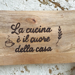 Könnte beinhalten: Holzschneidebrett mit dem italienischen Satz "La cucina è il cuore della casa" (Die Küche ist das Herz des Hauses) in dunkelbraunen Buchstaben. Eine Kaffeetasse und eine Weizenähre sind ebenfalls eingeätzt.