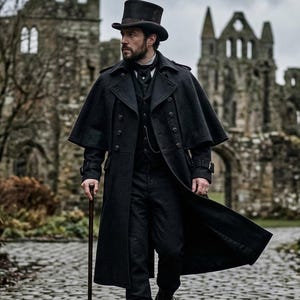 Pode incluir: Uma pessoa com um casaco preto, chapéu alto e botas, segurando uma bengala, caminha por um caminho de paralelepípedos. O casaco tem um design tipo capa. O fundo apresenta um edifício de pedra e um céu nublado.