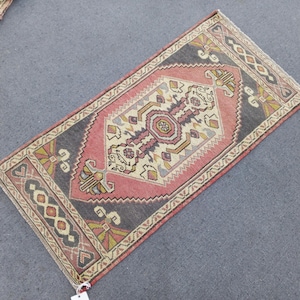 May include: A rectangular, hand-woven rug with a central medallion design. The rug features a pink and grey colour scheme with intricate geometric patterns in beige, yellow, and burgundy. The rug's border has a repeating pattern. The rug is approximately 91 cm long and 51 cm wide.