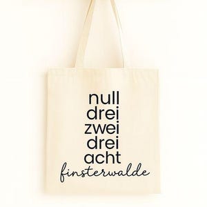 Könnte beinhalten: Cremefarbene Tragetasche mit schwarzem Text "null drei zwei drei acht finsterwalde". Die Tasche hängt an einer Holzgarderobe mit Stiften. Die Tasche hat ein schlichtes Design und ist wahrscheinlich aus Canvas oder einem ähnlichen Material.