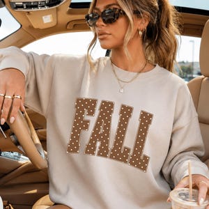 Könnte beinhalten: Ein sandfarbenes Sweatshirt mit Rundhalsausschnitt und dem Wort FALL, das in großen, braunen Blockbuchstaben mit weißem Punktmuster aufgedruckt ist. Das Sweatshirt hat lange Ärmel, einen gerippten Halsausschnitt und eine lockere Passform in Beige.