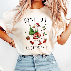May include: An ivory t-shirt featuring a graphic of a goose wearing a red and white Santa hat, a red patterned sweater, and red boots. The goose carries a green Christmas tree. Text reads OOPS! I GOT ANOTHER TREE with candy cane and gift icons.