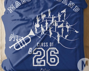 Può includere: Una coperta blu con le scritte "MARCHING BAND" e "CLASS OF '26" in bianco. Sono presenti anche una tromba, note musicali e figure di banda musicale. La coperta è su un letto con cuscini bianchi.