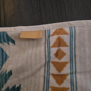 Puede incluir: Primer plano de una toalla estampada beige y turquesa. La toalla presenta un dise&ntilde;o geom&eacute;trico con tri&aacute;ngulos y diamantes en tonos marrones y turquesas. Una peque&ntilde;a etiqueta rectangular de color canela est&aacute; cosida en el borde.