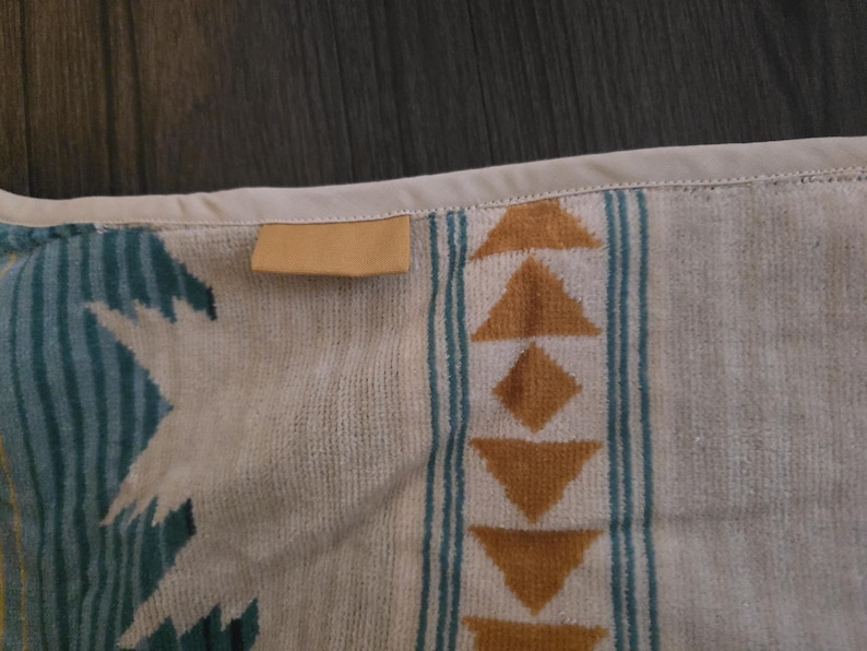 Puede incluir: Primer plano de una toalla estampada beige y turquesa. La toalla presenta un dise&ntilde;o geom&eacute;trico con tri&aacute;ngulos y diamantes en tonos marrones y turquesas. Una peque&ntilde;a etiqueta rectangular de color canela est&aacute; cosida en el borde.