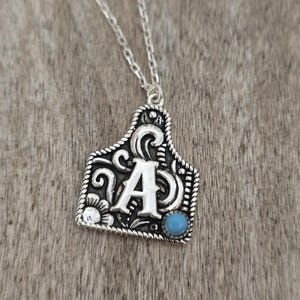Op de afbeelding: Zilverkleurige hangerketting met een rechthoekig tag-ontwerp met de letter "A" in het midden. De hanger heeft decoratieve krullen, een kleine turkooizen steen en een touwachtige rand. De ketting ligt op een houten oppervlak.