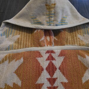 Puede incluir: Una toalla con capucha con un patr&oacute;n geom&eacute;trico en tonos naranja, blanco y azul. La toalla est&aacute; hecha de un material suave y absorbente y tiene una capucha para mayor calidez y comodidad. El dise&ntilde;o presenta un patr&oacute;n repetido de diamantes y tri&aacute;ngulos.