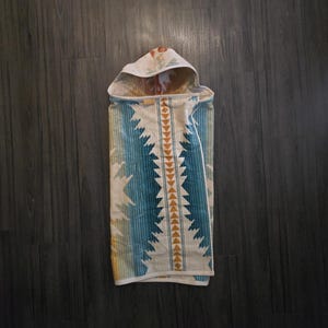 Puede incluir: Una toalla con capucha para beb&eacute; con un dise&ntilde;o de inspiraci&oacute;n suroeste. La toalla presenta un patr&oacute;n de rayas y tri&aacute;ngulos azules, blancos y amarillos. La capucha tiene un estampado floral. La toalla est&aacute; doblada sobre una superficie de madera oscura.