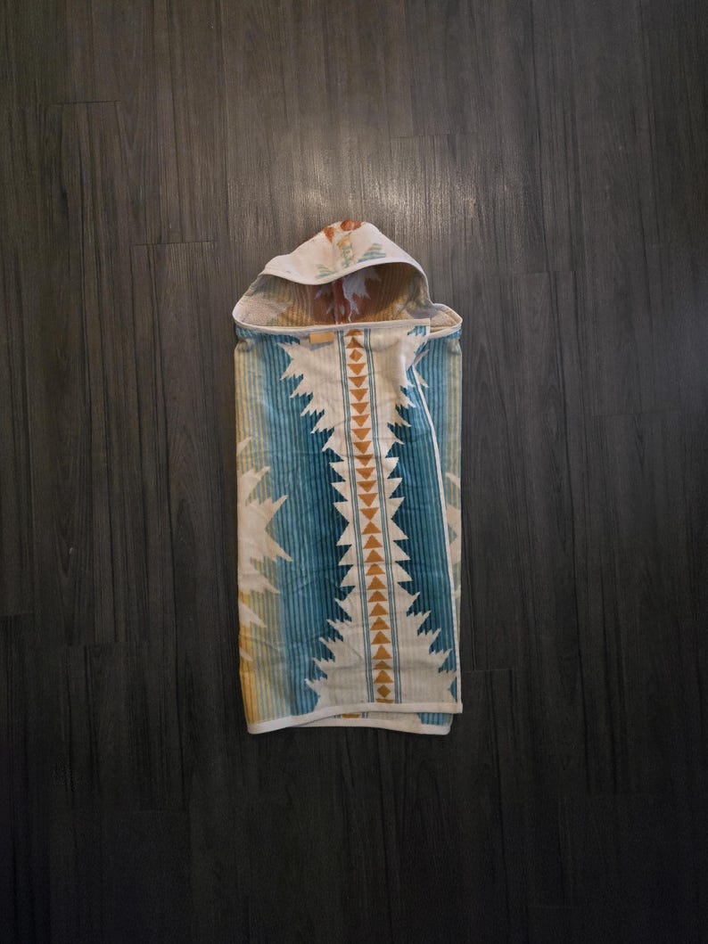 Puede incluir: Una toalla con capucha para beb&eacute; con un dise&ntilde;o de inspiraci&oacute;n suroeste. La toalla presenta un patr&oacute;n de rayas y tri&aacute;ngulos azules, blancos y amarillos. La capucha tiene un estampado floral. La toalla est&aacute; doblada sobre una superficie de madera oscura.